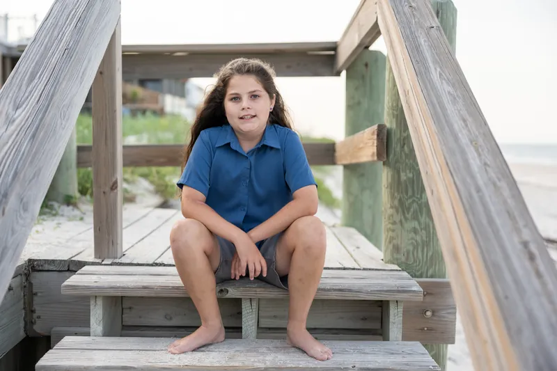 Girl in blue polo shirt sits barefoot on wooden bench structure, gazing at camera.