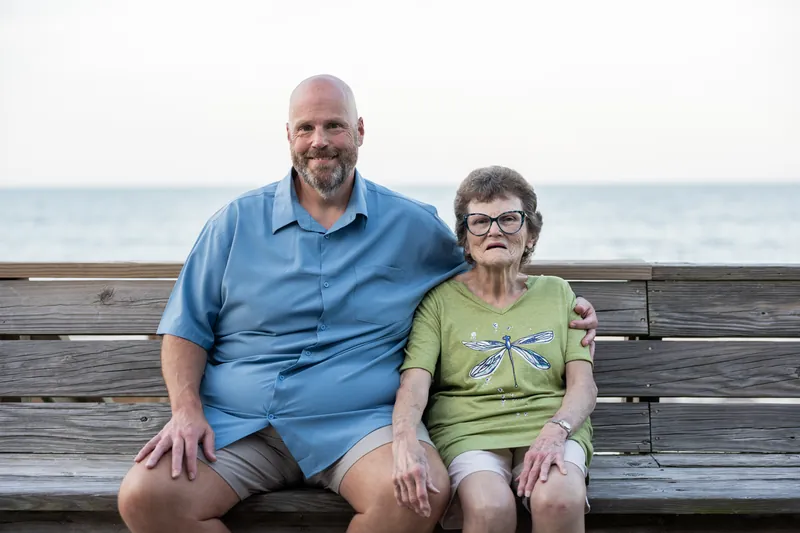 Man in blue shirt and woman in lime green dragonfly shirt sit together on wooden bench at St. Augustine Beach.