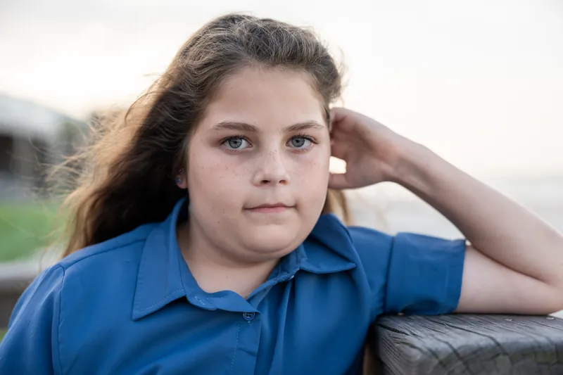 Girl in blue polo shirt rests her head on her hand with a calm expression outdoors.