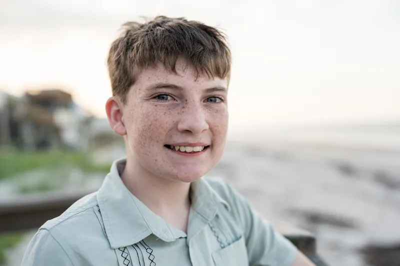 Smiling boy with freckles wearing a sage button-up shirt against an overcast sky.