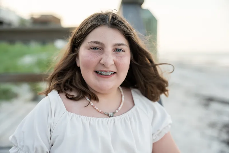Girl in a white short-sleeved top with long brown hair smiling at the camera by the water.