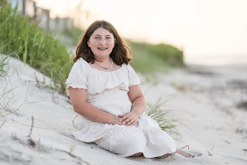 Young girl in white sitting on sand dunes at St. Augustine Beach, hands folded in her lap.