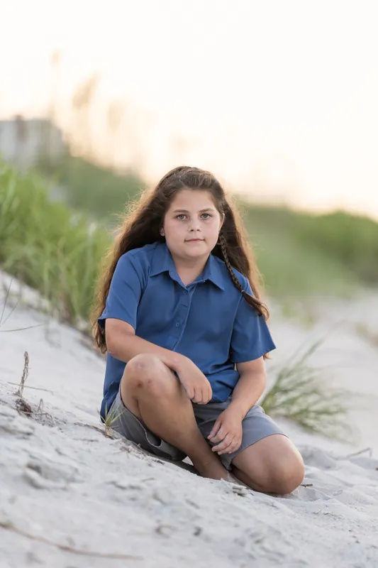 Preteen girl in navy kneeling on sandy beach at St. Augustine Beach with sea grass behind her.