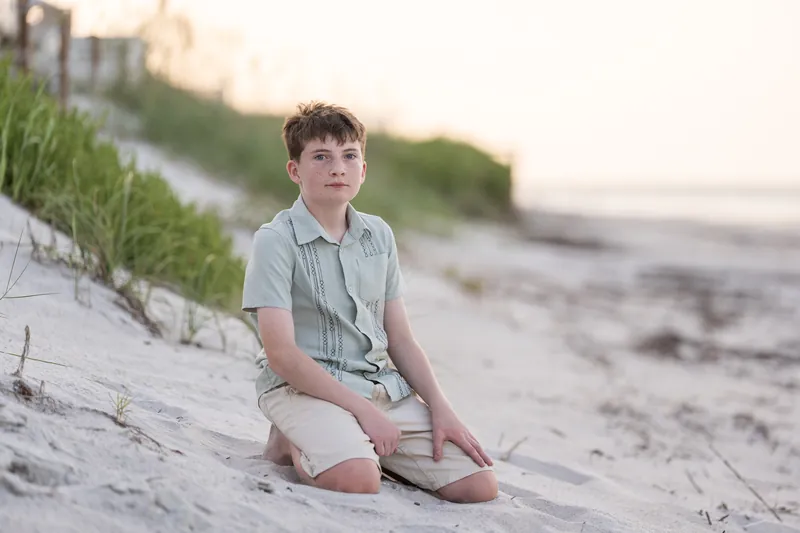 Boy in light gray shirt seated on sand at St. Augustine Beach, looking toward the camera.