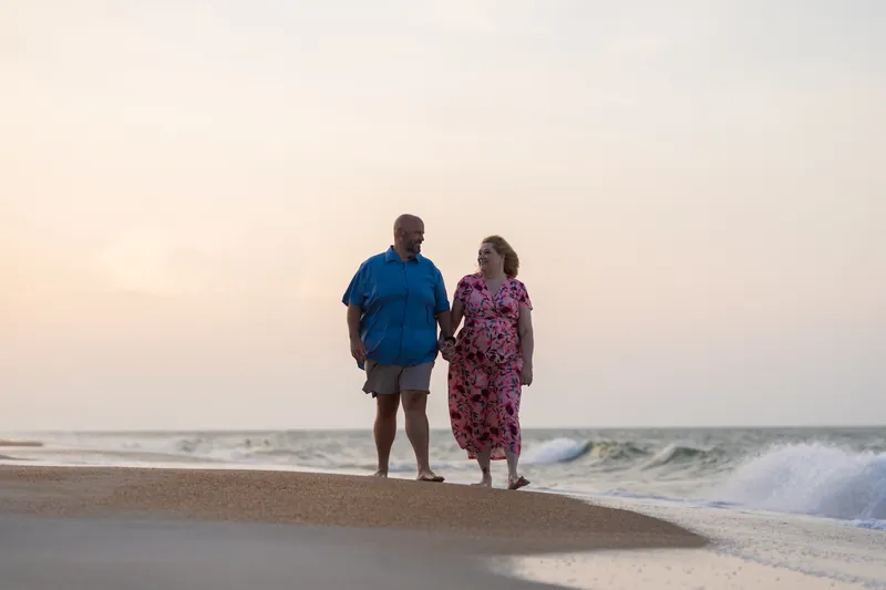 A man in a blue shirt and woman in a floral dress stand ankle-deep in the ocean at St. Augustine Beach at sunset.