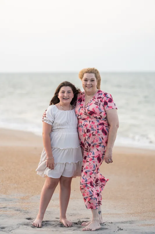 Two girls smile at the camera while standing on the beach at St. Augustine, one wearing a white tiered dress and the other in pink floral.