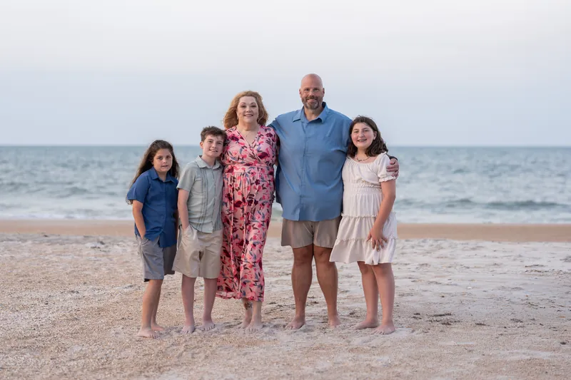 Trena's family of five poses together on the sand at St. Augustine Beach with the ocean and sky behind them.