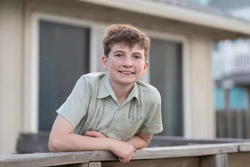 Boy in a sage button-up shirt leans against a wooden railing, smiling directly at the camera.