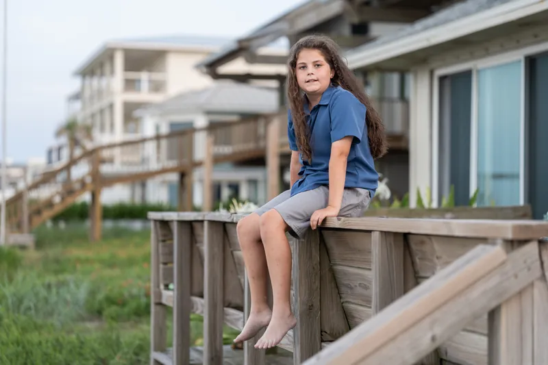 Girl in a blue shirt sits on a wooden deck railing with her legs dangling, looking over her shoulder.