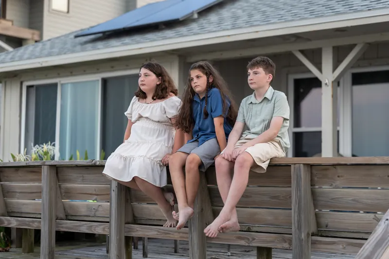 Three children perch barefoot on a wooden deck railing—a girl in white on the left, girl in blue in the middle, boy in sage on the right.