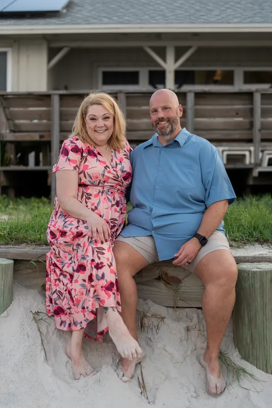 Trena and her husband sit together on a weathered wooden bench, both wearing short-sleeved shirts and smiling at the camera.