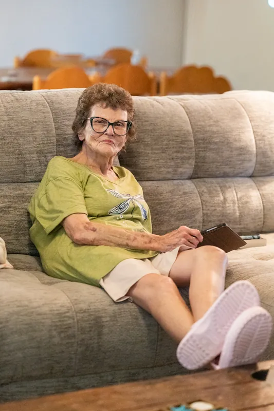 Trena sits on a beige sofa wearing a lime-green shirt and lavender socks, smiling at the camera with her arms relaxed.