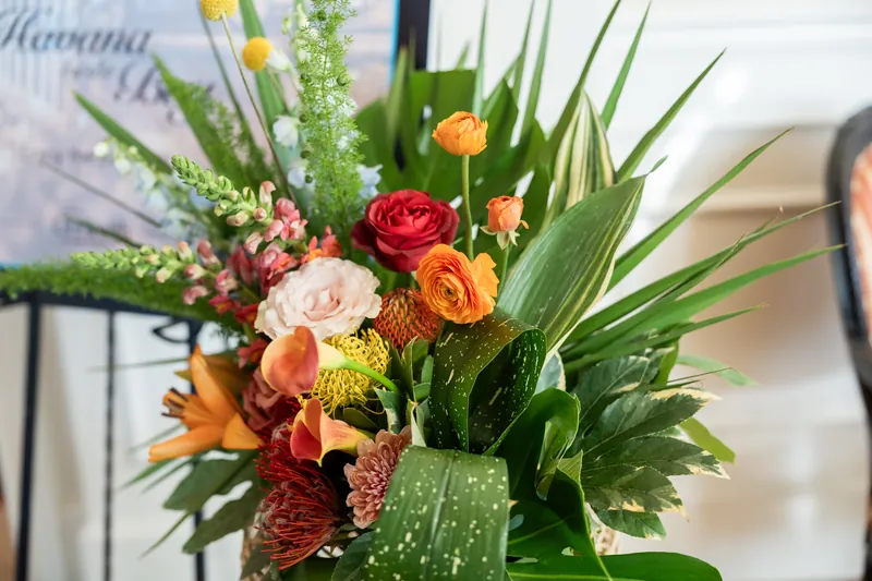Close-up of lush mixed bouquet with red, coral, and blush roses, green foliage, and tropical leaves with water droplets.