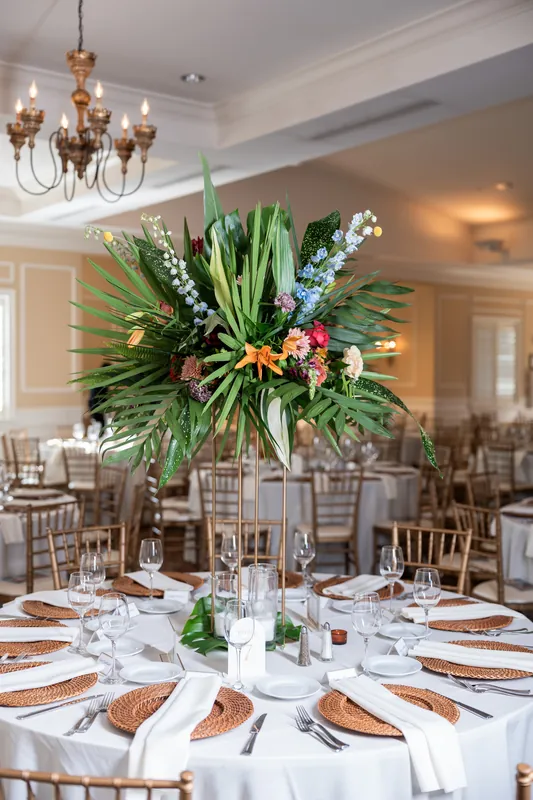 Round dining table set with white linens, gold-rimmed plates, and tall tropical floral centerpiece under crystal chandelier at River House Events.