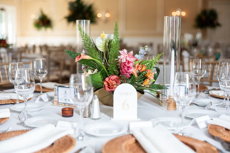 Intimate table setting with white linens, place card, and mixed floral arrangement in clear cylinder vase with green foliage and coral blooms.