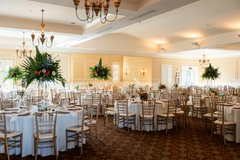 Formal dining room at River House Events features round tables with blue linens, chiavari chairs, and hanging greenery-filled spheres beneath brass chandeliers.