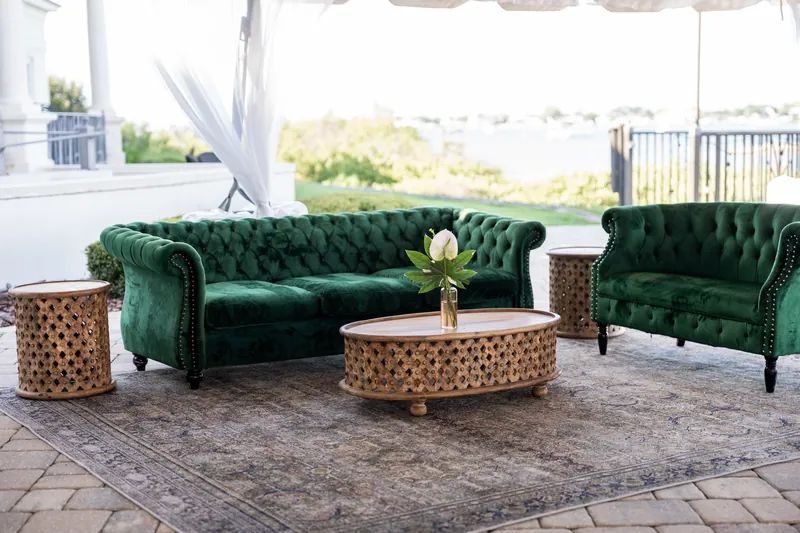 Green tufted sofa and armchair with woven ottomans sit on a patterned rug under a white tent at River House Events.