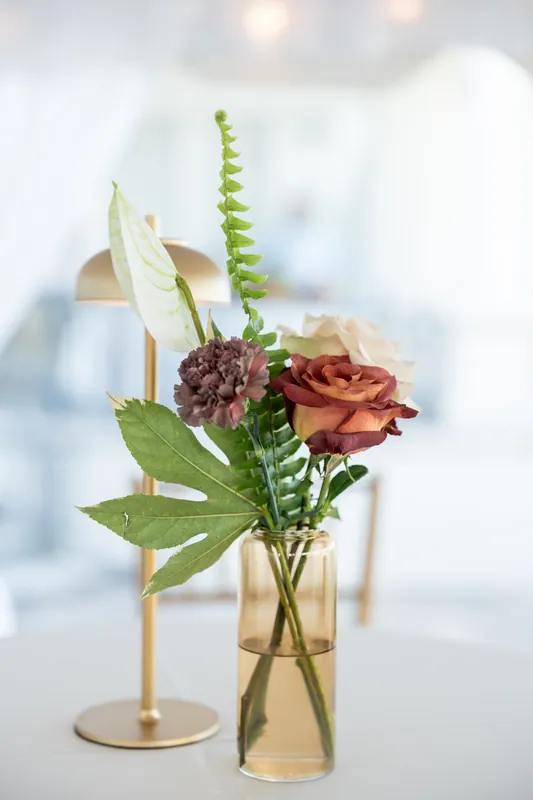 Tall glass vase holds mauve and rust-toned flowers with cascading green fern and white foliage.