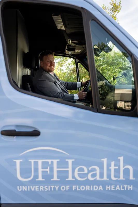 A person in a dark suit sits in the driver's seat of a blue UF Health van, visible through the open window.