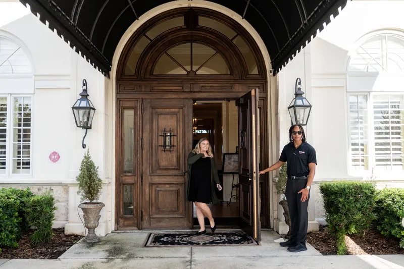 Two staff members flank the arched wooden entrance door as a guest in a dark dress arrives at River House Events.