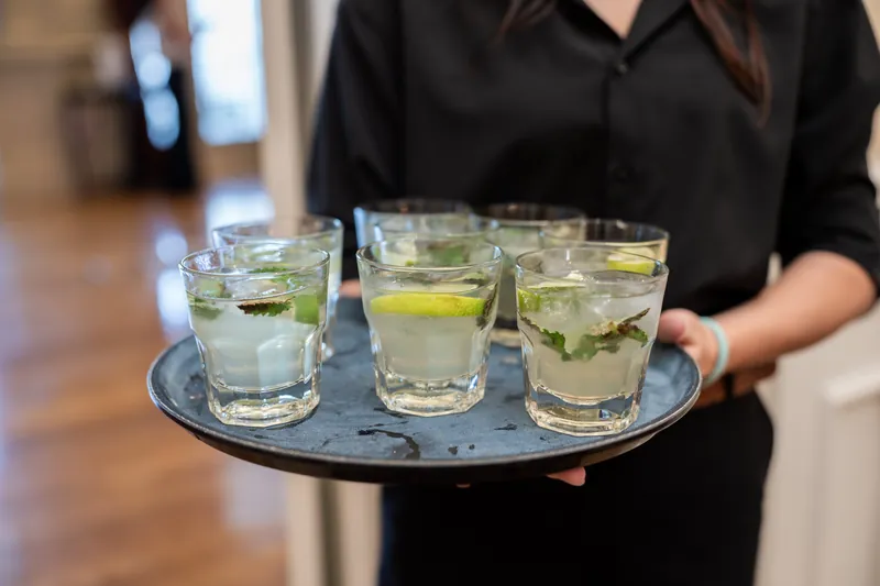 Server in black uniform holds a silver tray with three glasses of water and lime garnish.