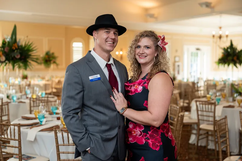 Two people in formal attire smile together in the dining room, one wearing a dark suit and hat, the other in a floral dress.