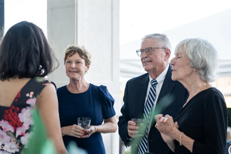 Four guests in business casual clothing hold drinks and converse near large windows with water views.