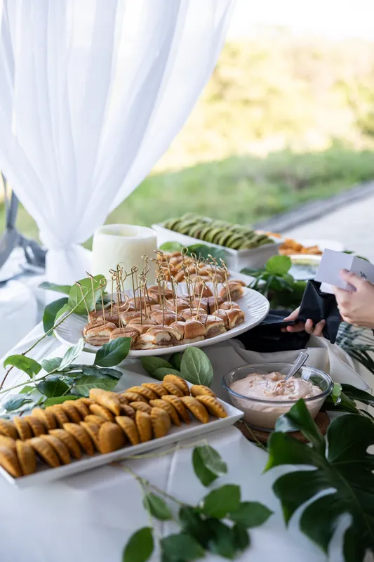 Appetizers and charcuterie spread across white linens under sheer draping with waterfront views in soft daylight.