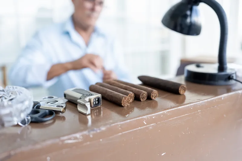 An array of cigars and smoking accessories displayed on a desk with a desk lamp and blurred person in background.
