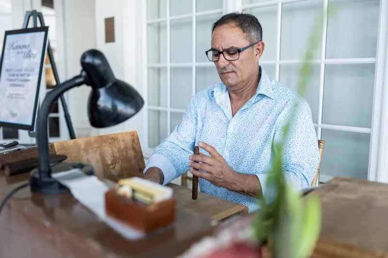 A man in a light blue shirt speaks with hands gesturing at a wooden table near a desk lamp.