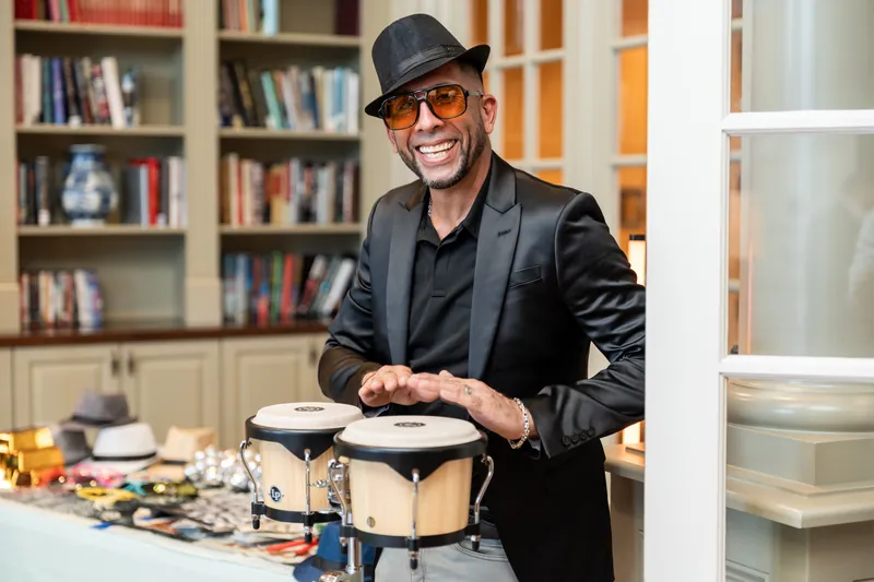 A man in a black blazer and hat plays a small drum at a table with art supplies nearby.
