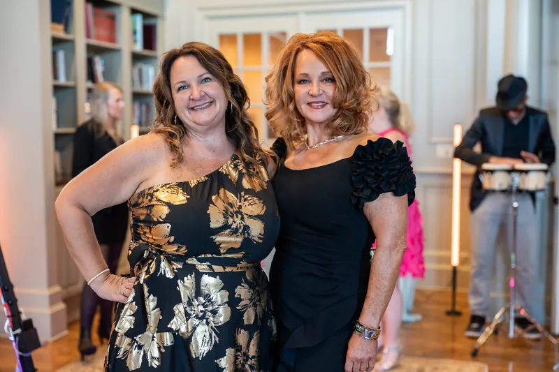 Two women in formal attire pose together at River House Events, with bookshelves and event staff visible behind them.