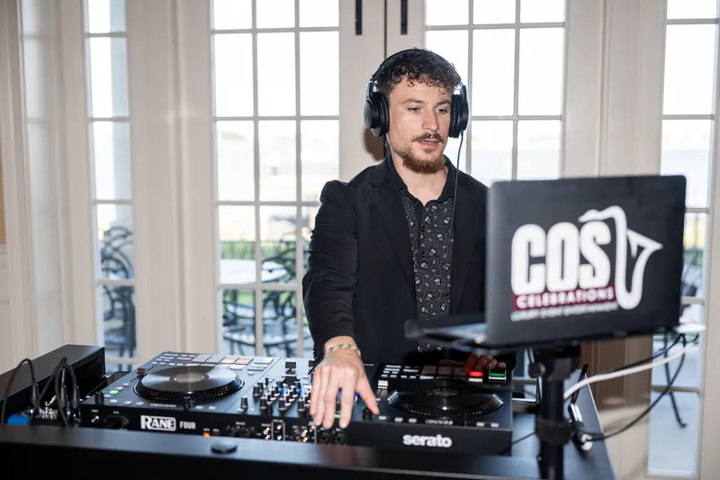 DJ in a black blazer and patterned shirt operates turntables with large windows overlooking the grounds behind him.