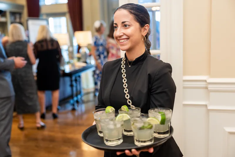 Server carrying a tray of lime-garnished cocktails moves through the crowded event space with a warm smile.