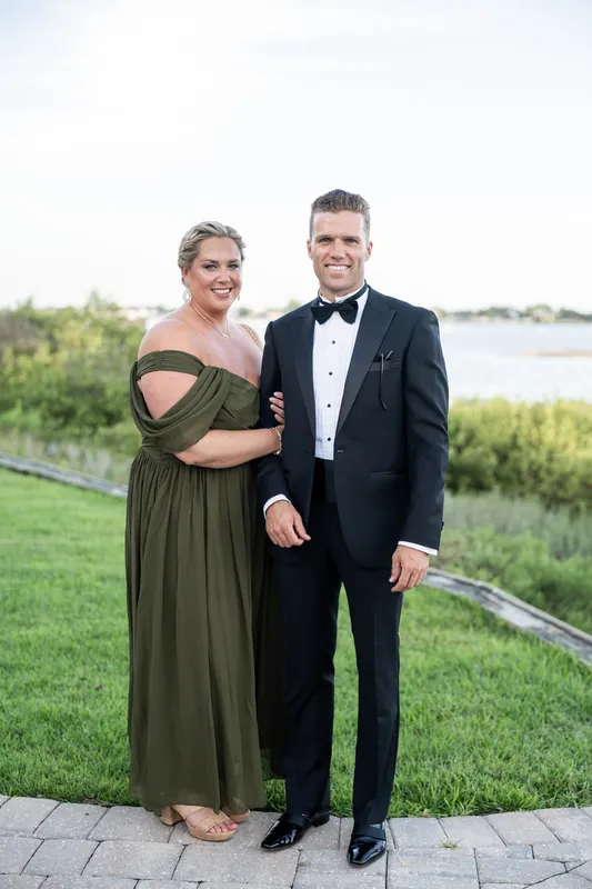 A pair in an olive gown and black tuxedo stand on a stone path with the waterway beyond.