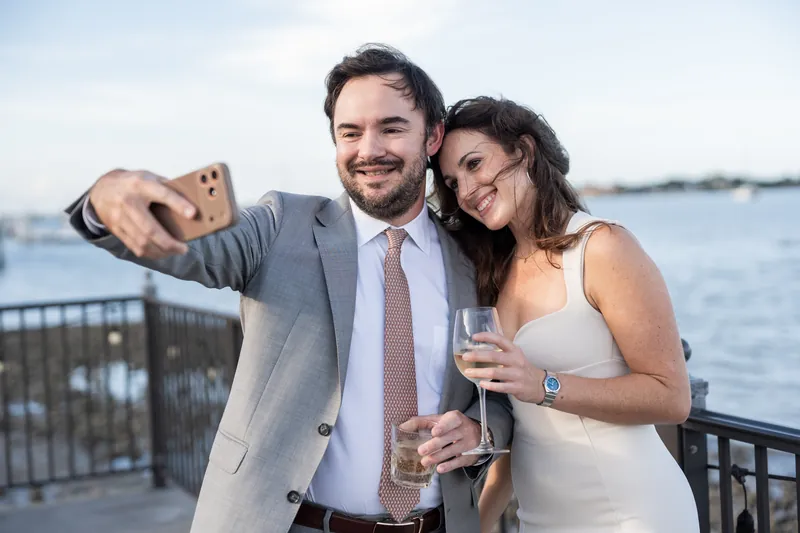 Two guests clink glasses and smile on the deck, one in a light blazer and mauve tie, the other in white.