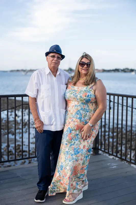 A man in a blue cap and white shirt stands with a woman in a colorful floral dress by the waterfront railing.