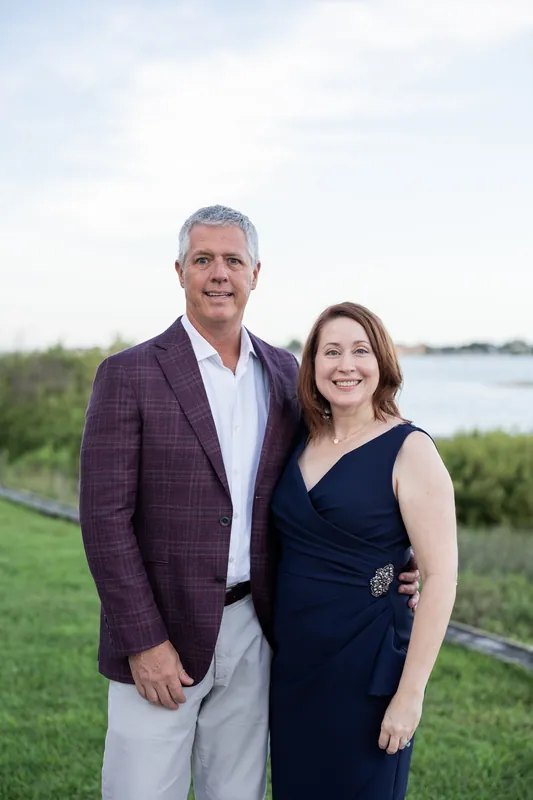 Two people smile for the camera on the lawn—one in a burgundy blazer, the other in a navy wraparound top.