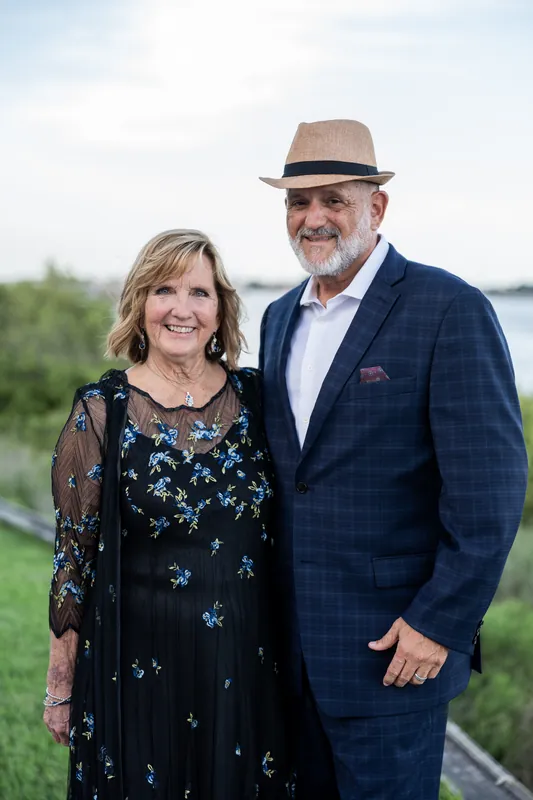 A couple poses together; she wears a navy embellished dress, he wears a navy plaid jacket and tan hat.