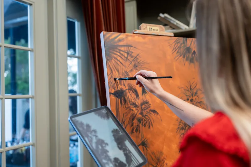 Someone in a bright red sleeve brushes dark leafy silhouettes onto an orange canvas near tall windows.