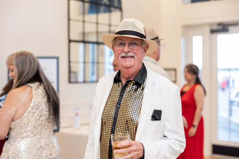 A man in a white jacket and Panama hat holds a drink at the indoor reception venue.
