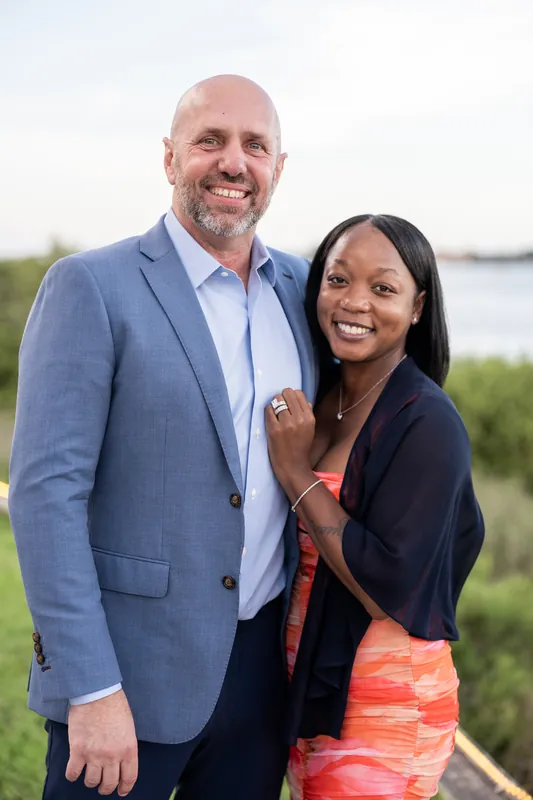 Two people pose together outdoors with the river visible in the background.