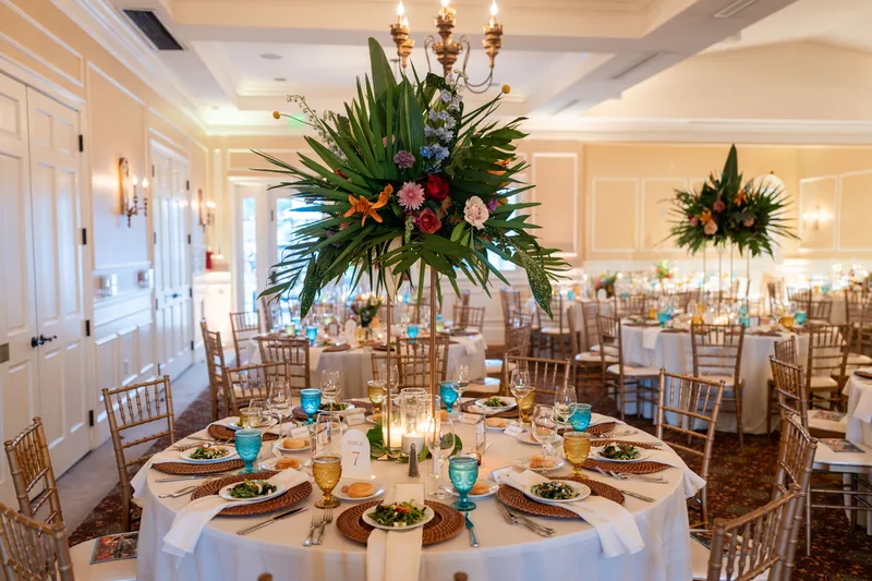 Round dining tables with tall floral centerpieces and turquoise accents fill the elegant ballroom.