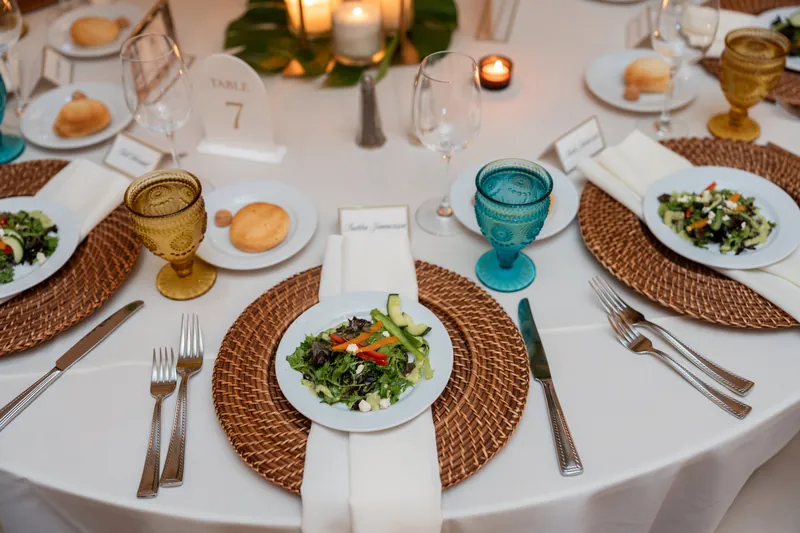 Overhead view of round table with salads on woven placemats, turquoise glassware, and table number cards at dinner.