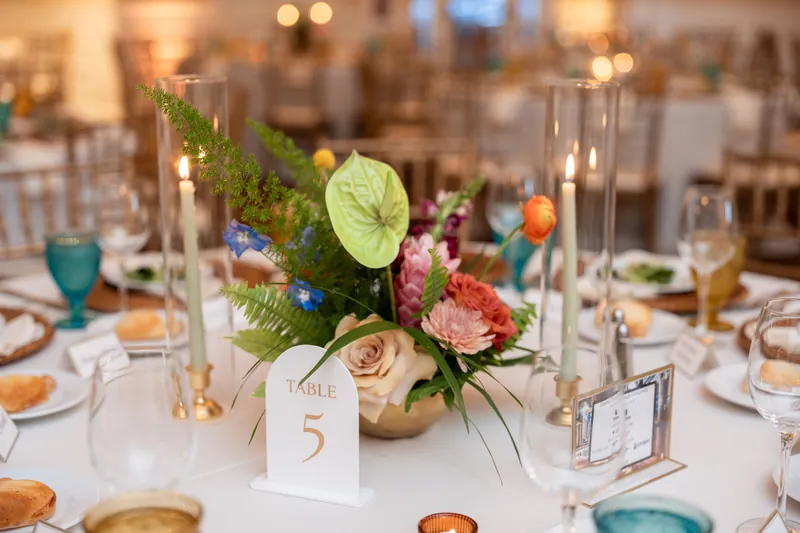 Floral centerpiece with roses, greenery, and a white "Table 5" card surrounded by place settings and candlelight.