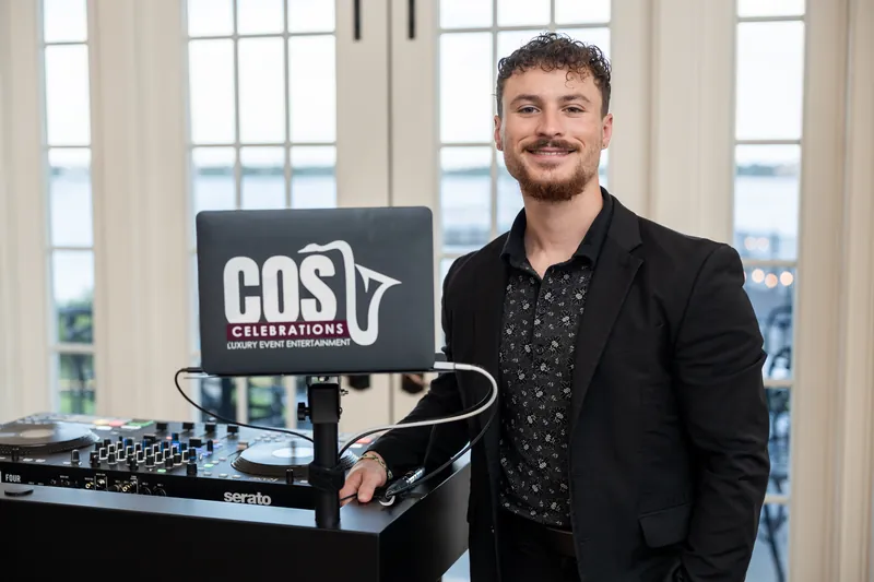 DJ in black blazer and patterned shirt stands at equipment with COSV Entertainment sign and water views behind.