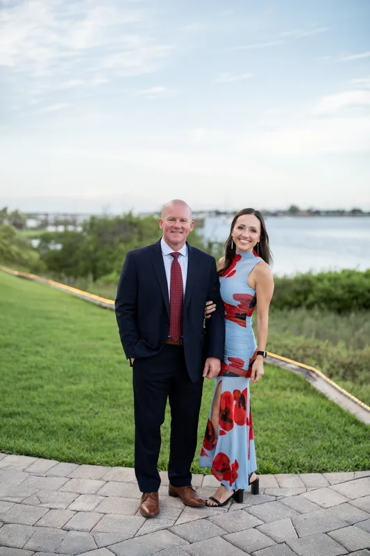 Two guests smile together on a manicured lawn with river views and ornamental railings at the venue's waterfront edge.