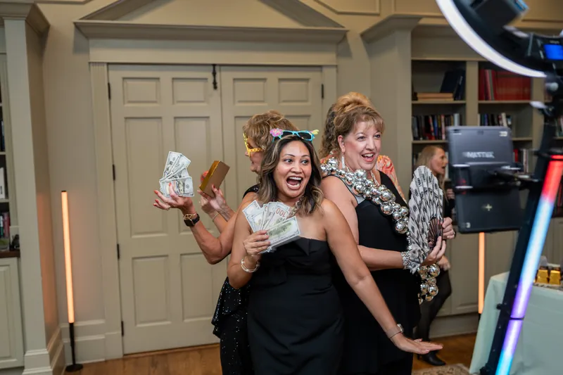 Three people laugh and hold cash near a photo booth setup in an indoor room with white cabinetry and wooden floors.