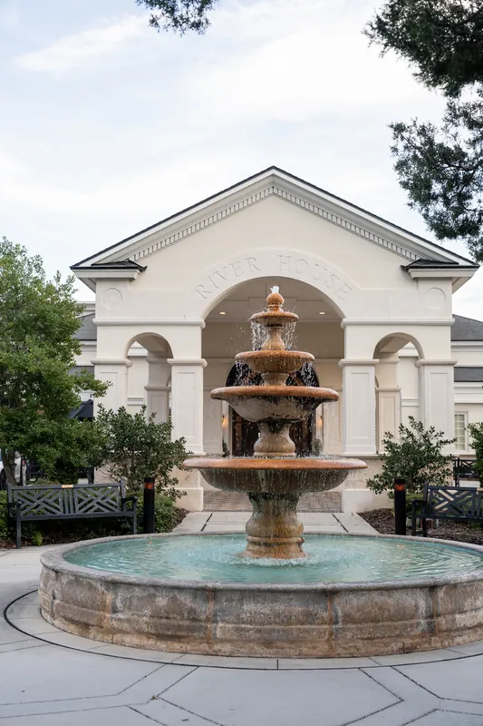 Tiered fountain anchors the courtyard at River House Events, with the cream and arched stone building framed by green landscaping under a pale sky.