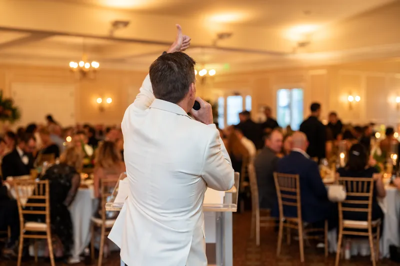 Someone in a white shirt addresses a crowded ballroom, turning toward guests with one hand raised.